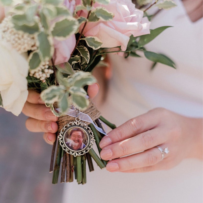 May include: A white bridal bouquet with pink roses and greenery, featuring a silver locket charm with a photo of a person inside.