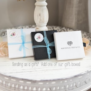 May include: Three square gift boxes, two white and one black, each tied with a light blue ribbon. The boxes feature a circular sticker with a floral design and the text Magnolia Tree & Co. The text Sending as a gift? Add one of our gift boxes! is visible.