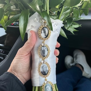 May include: A wedding bouquet wrapped in white satin ribbon with scattered clear crystals, featuring a vertical gold-toned metal charm strand with four oval frames containing black and white portrait photographs of individuals.