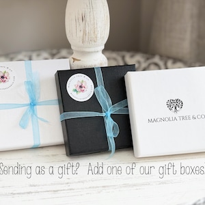 May include: Three gift boxes: white, black, and white. Each box is tied with a light blue ribbon. The white boxes have a floral sticker and the text "MAGNOLIA TREE & CO." The image includes the text "Sending as a gift? Add one of our gift boxes!"