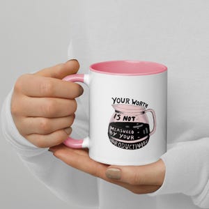 May include: White ceramic mug with a pink interior and handle. The mug features a graphic of a coffee pot with the text "YOUR WORTH IS NOT MEASURED BY YOUR PRODUCTIVITY". The mug is held by a person.