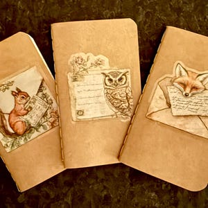 May include: Three beige notebooks with animal illustrations. One notebook features a squirrel holding a letter, another an owl, and the third a fox with a letter. The notebooks have a vintage aesthetic.