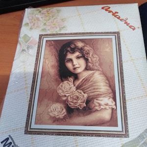 Op de afbeelding: Een kruissteekpatroon met een portret van een vrouw met bruin haar en een sjaal, die een boeket rozen vasthoudt. Het patroon is ingelijst in een bruin frame en heeft de tekst "Ariadna" in de rechterbovenhoek.