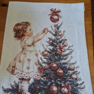 May include: A decorative Christmas-themed tapestry featuring a young girl decorating a Christmas tree. The girl is wearing a dress with snowflake patterns. The tree is adorned with red ornaments and bows. The background is a light, textured fabric.