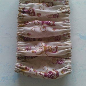 以下が含まれることがあります： 白地に茶色とピンクのフクロウ柄がプリントされた布製のリストレット。リストレットは上部がギャザーになっており、白い紐が付いています。