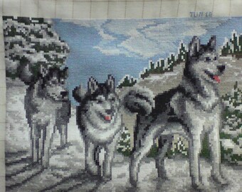May include: A cross-stitch embroidery of three gray and white wolves in a snowy landscape. The wolves are standing in a line, with the largest wolf in the back and the smallest wolf in the front. The wolves are all looking to the right, and the background is a snowy forest.