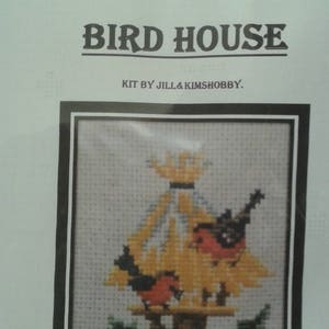 May include: A cross-stitch kit depicting a birdhouse with two red birds perched on the edge. The birdhouse is yellow with a brown roof and is surrounded by green leaves and red berries. The kit is titled "Bird House" and includes the text "Kit by Jill & Kimshobby."