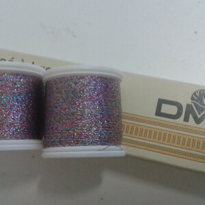 Può includere: Due bobine di filo metallico DMC, una rosa e una blu, con un luccichio argentato. Le bobine sono etichettate "Fil métallique" e "DMC".