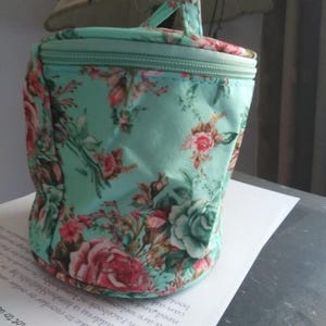May include: A small, rounded cosmetic bag with a handle, featuring a teal background and a floral pattern of pink and red roses with green leaves. The bag has a zipper closure and a matching teal handle.