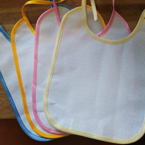 May include: Four white bibs with different colored trim. The bibs are made of a soft, mesh-like material and have a rounded neckline. The trim colors are blue, yellow, pink, and cream.