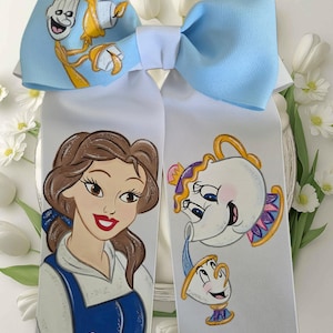 May include: A light blue and white hair bow featuring a printed illustration of Belle, Mrs. Potts, Chip, and Lumiere from Beauty and the Beast. The bottom of the white ribbon tails displays the black text Hugs And Kisses Boutique.