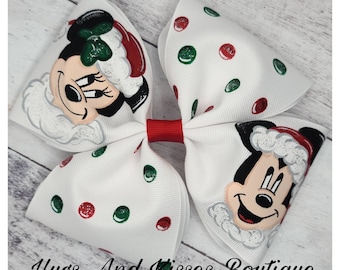 Puede incluir: Un lazo blanco grande con lunares rojos y verdes y un centro rojo. El lazo presenta ilustraciones de Mickey y Minnie Mouse con gorros de Papá Noel. El texto "Hugs And Kisses Boutique" está en la parte inferior.