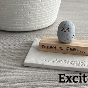 Thumb-sized rock shaped figurine with excited emoji expression sitting on a mini wooden plank with the phrase "today I feel"