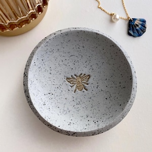 K&ouml;nnte beinhalten: Eine kleine, graue, gesprenkelte, runde Schale mit einem goldenen Bienen-Design. Die Schale ist perfekt zum Aufbewahren von Schmuck oder anderen kleinen Gegenst&auml;nden.
