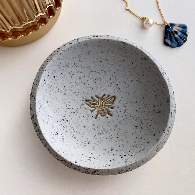 K&ouml;nnte beinhalten: Eine kleine, graue, gesprenkelte, runde Schale mit einem goldenen Bienen-Design. Die Schale ist perfekt zum Aufbewahren von Schmuck oder anderen kleinen Gegenst&auml;nden.