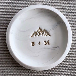 Ring dish with White and accents silver gray resembling marble. Stamped with a mountain design in gold and couple's initials below the mountain. For example, B+M