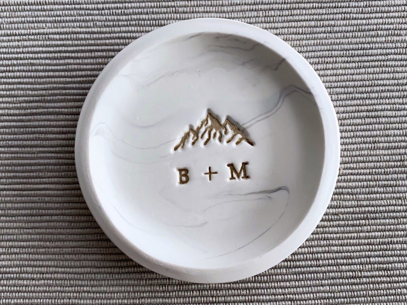 Ring dish with White and accents silver gray resembling marble. Stamped with a mountain design in gold and couple's initials below the mountain. For example, B+M