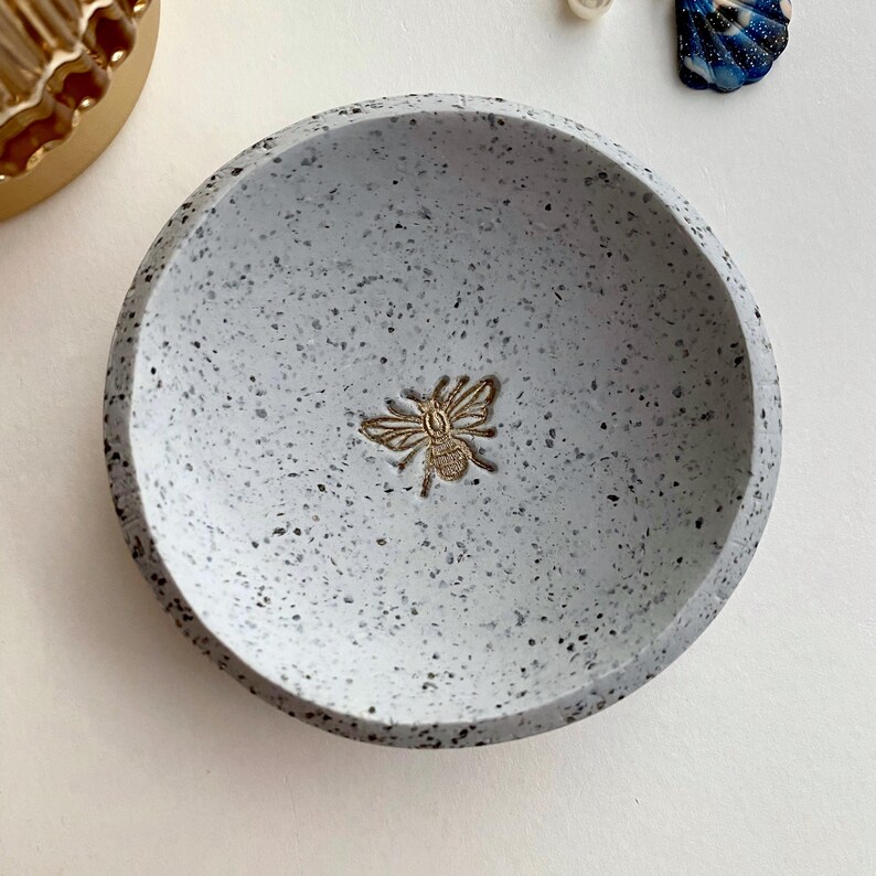K&ouml;nnte beinhalten: Eine kleine, wei&szlig; gesprenkelte Keramikschale mit einem goldenen Bienen-Design. Die Schale ist perfekt f&uuml;r die Aufbewahrung von Schmuck oder anderen kleinen Gegenst&auml;nden.