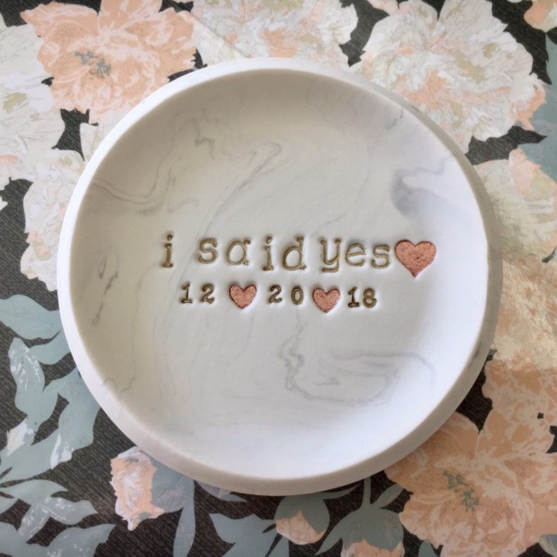 May include: White ceramic ring dish with a marble pattern. It says "i said yes" with a gold heart and "12 20 18" with three rose gold hearts.