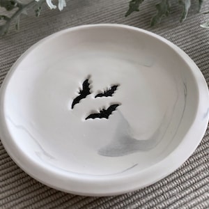 May include: A white ceramic dish with a marbled design and three black bat cutouts. The dish is perfect for holding jewelry or other small items.