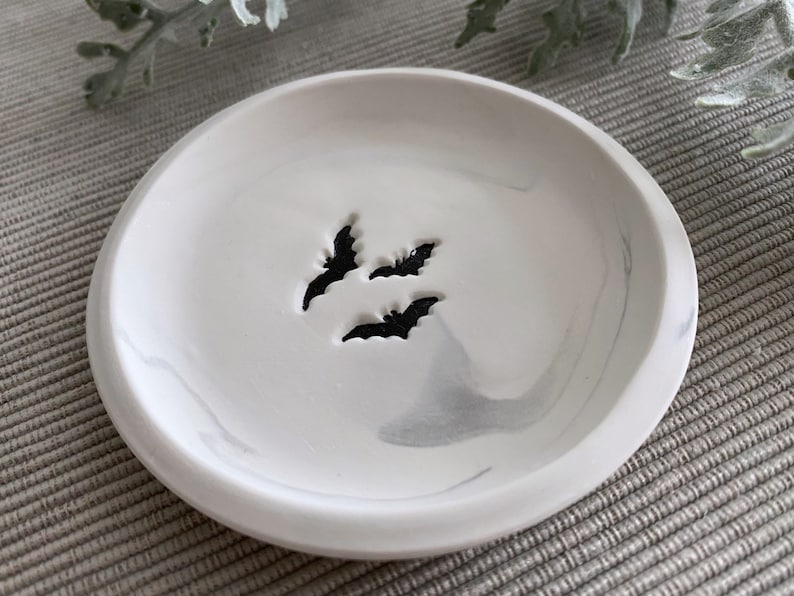 May include: A white ceramic dish with a marbled design and three black bat cutouts. The dish is perfect for holding jewelry or other small items.
