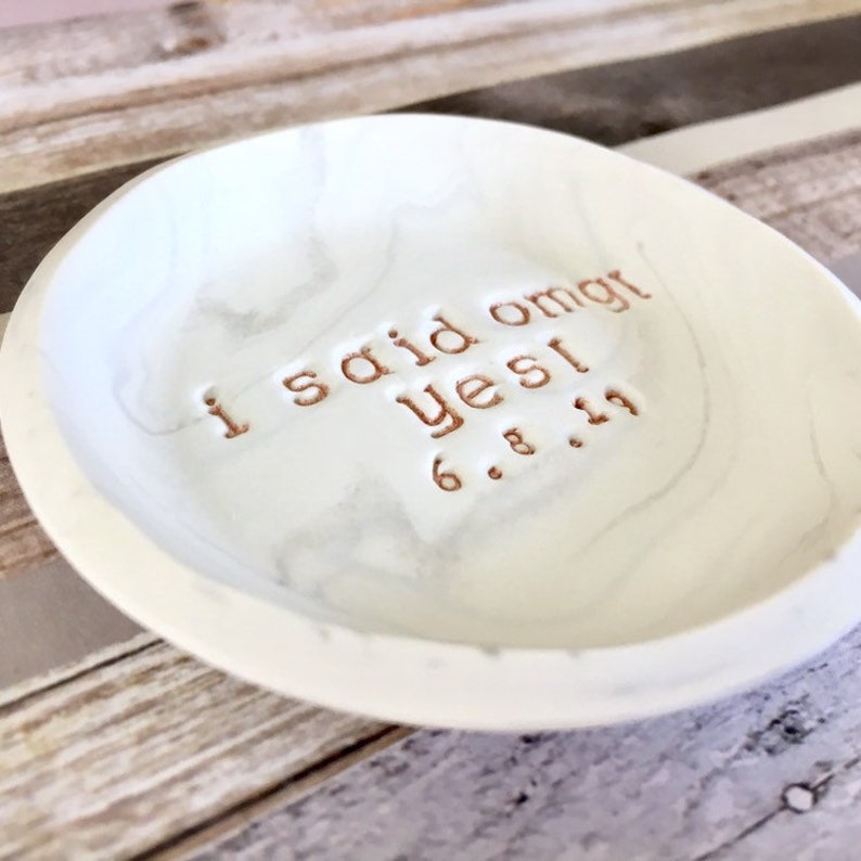 May include: White ceramic ring dish with a marble pattern. The dish has the words "I said omg yes! 6.8.19" inscribed in gold lettering.
