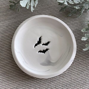 May include: A white ceramic dish with a marbled design and three bat silhouettes. The dish is perfect for holding jewelry, keys, or other small items.