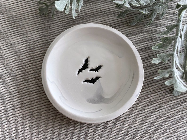 May include: A white ceramic dish with a marbled design and three bat silhouettes. The dish is perfect for holding jewelry, keys, or other small items.