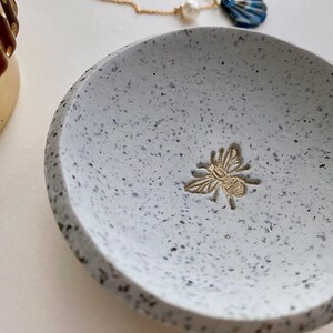 K&ouml;nnte beinhalten: Eine kleine, graue, gesprenkelte Keramikschale mit einem goldenen Bienen-Design. Die Schale ist perfekt zum Aufbewahren von Schmuck oder anderen kleinen Gegenst&auml;nden.