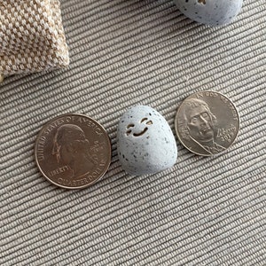 Thumb-sized rock shaped figurine with happy emoji expression in between a quarter and nickel for size comparison
