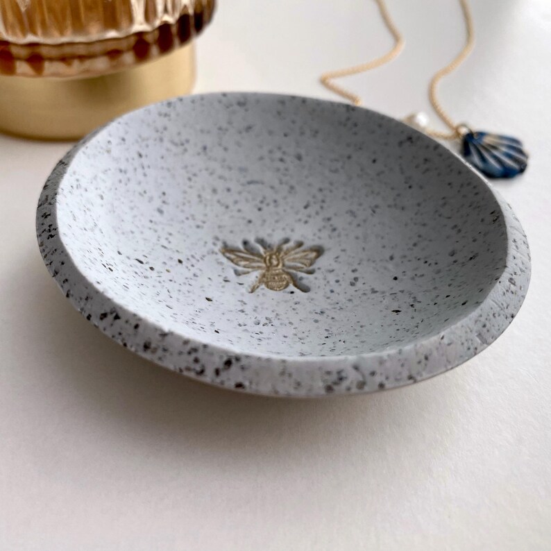 K&ouml;nnte beinhalten: Eine kleine, wei&szlig;e Keramikschale mit gesprenkeltem Finish. Die Schale hat ein erhabenes Bienendesign in der Mitte.