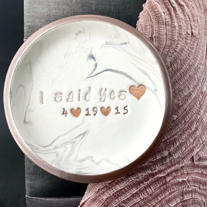 May include: White ceramic ring dish with a marble pattern and a brown rim. The dish has the words "i said yes" and the date "4 19 15" inscribed in cursive with small hearts between the numbers.