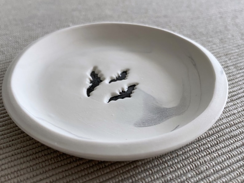 May include: A white ceramic trinket dish with a gray marbled design and a black tree silhouette in the center.