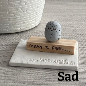 Thumb-sized rock shaped figurine with sad emoji expression sitting on a mini wooden plank with the phrase "today I feel"