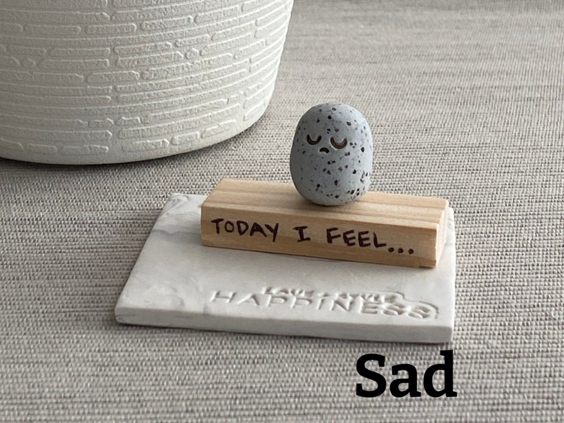 Thumb-sized rock shaped figurine with sad emoji expression sitting on a mini wooden plank with the phrase "today I feel"