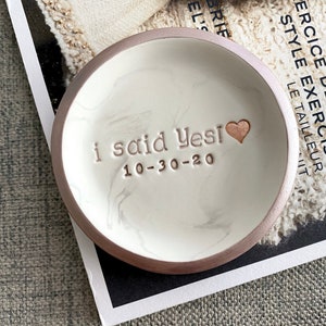 May include: A white ceramic ring dish with a marbled design and a gold rim. The dish has the words "i said Yesi 10-30-20" inscribed on it.