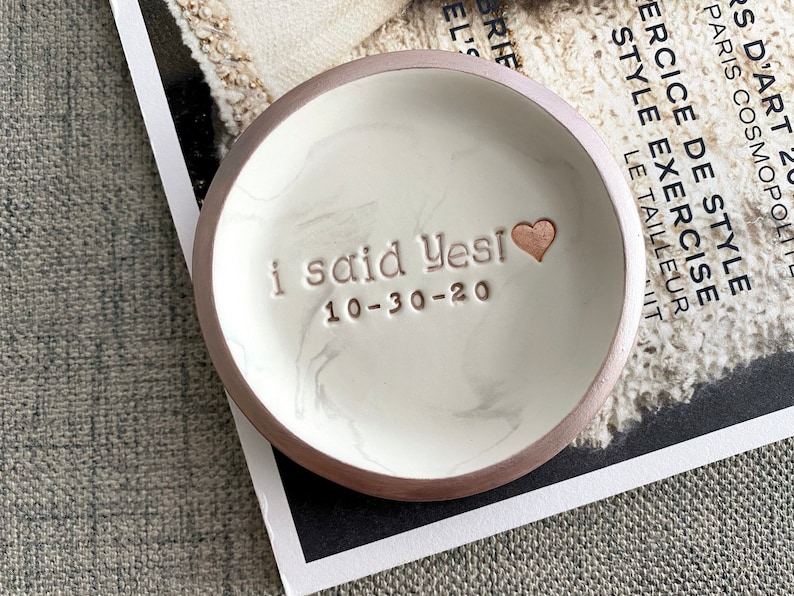 May include: A white ceramic ring dish with a marbled design and a gold rim. The dish has the words "i said Yesi 10-30-20" inscribed on it.
