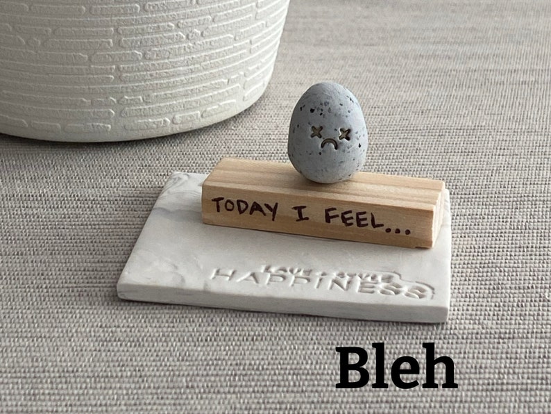Thumb-sized rock shaped figurine with x's for eyes emoji expression sitting on a mini wooden plank with the phrase "today I feel"