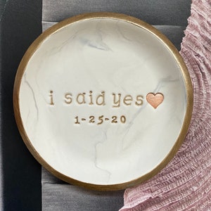 May include: White ceramic ring dish with a gold rim and a marble pattern. The dish has the words "i said yes" and the date "1-25-20" inscribed in gold lettering. A small gold heart is next to the text.