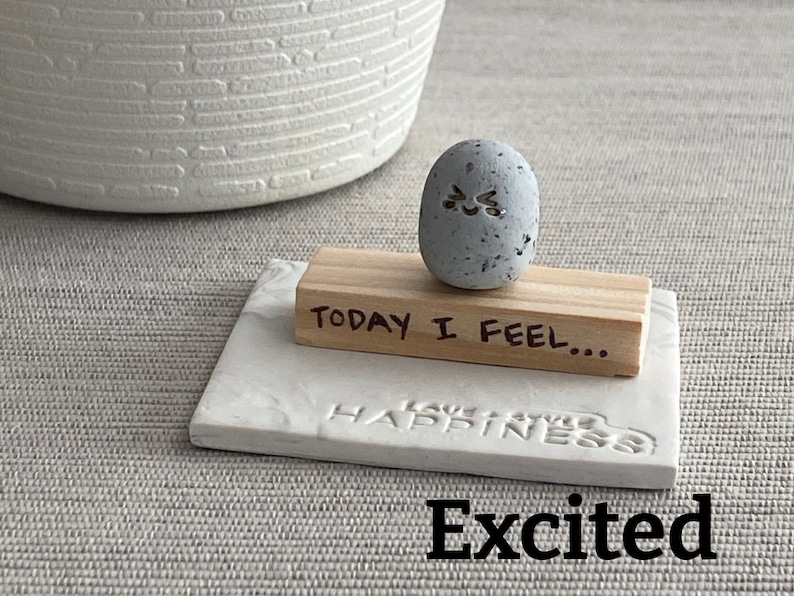 Thumb-sized rock shaped figurine with excited emoji expression sitting on a mini wooden plank with the phrase "today I feel"