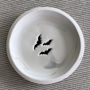 May include: A white ceramic dish with a marbled design and three bat cutouts. The dish is perfect for holding jewelry or other small items.