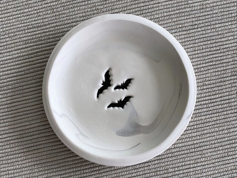 May include: A white ceramic dish with a marbled design and three bat cutouts. The dish is perfect for holding jewelry or other small items.