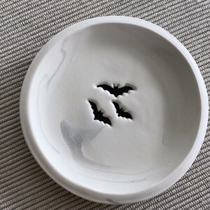 May include: A white ceramic dish with a gray marbled design. The dish has three bat silhouettes cut out of the center.