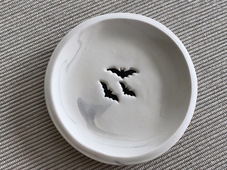 May include: A white ceramic dish with a gray marbled design. The dish has three bat silhouettes cut out of the center.