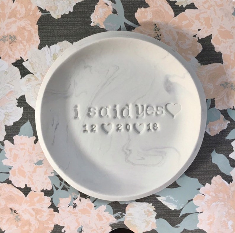 May include: White ceramic ring dish with a marbled design. The dish is engraved with the text "i said yes" and the date "12 20 18".