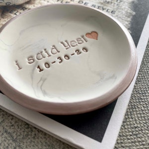 May include: White ceramic ring dish with a brown rim and a marbled design. The dish has the words "I said yes! 10-30-20" inscribed on it in gold lettering. A small gold heart is also inscribed on the dish.