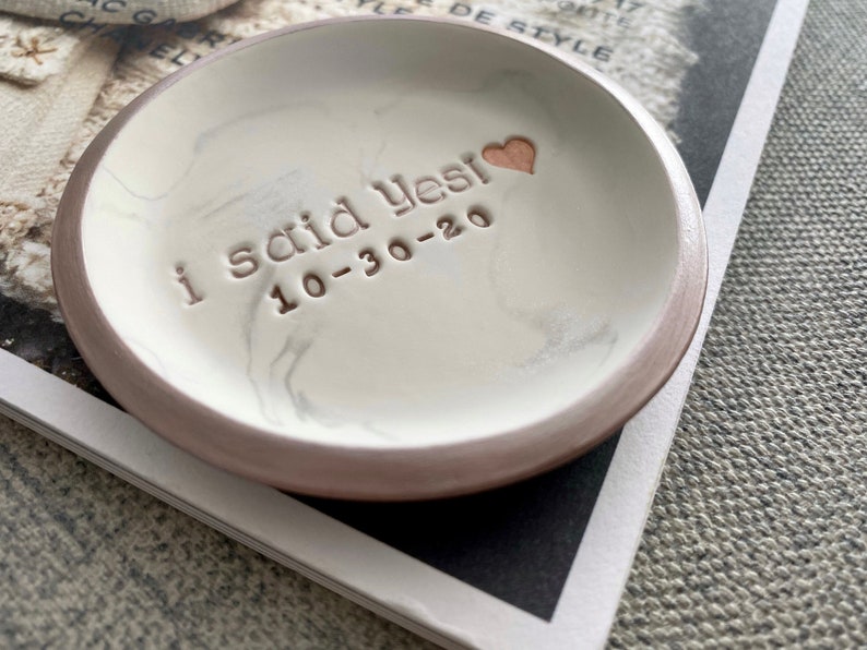 May include: White ceramic ring dish with a brown rim and a marbled design. The dish has the words "I said yes! 10-30-20" inscribed on it in gold lettering. A small gold heart is also inscribed on the dish.