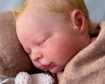 May include: A realistic newborn doll with pale skin, light blonde hair, and closed eyes, resting against a brown fabric surface. The doll is positioned next to a small, white, textured crocheted toy with black accents.