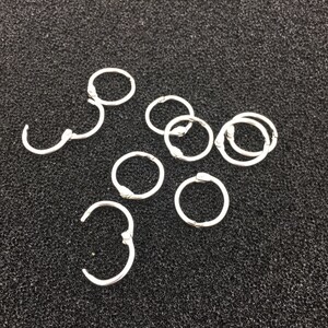 May include: A set of silver metal binder rings, also known as ring binders, for organising papers and documents. The rings are arranged in a scattered pattern on a dark grey textured surface.