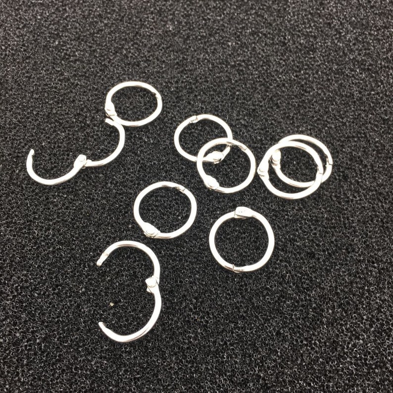 May include: A set of silver metal binder rings, also known as ring binders, for organising papers and documents. The rings are arranged in a scattered pattern on a dark grey textured surface.